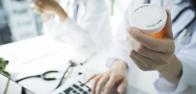 Physician holding up a pill bottle while working at a laptop